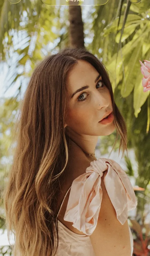 Elizabeth Faye under palm trees, hair down, a pink bow on her shoulder